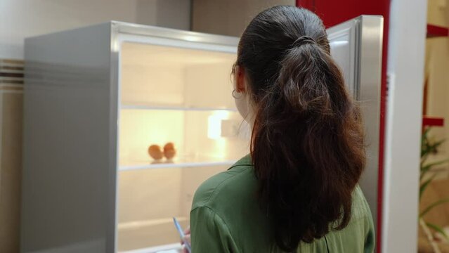 Young Caucasian woman opens empty refrigerator and writes down list of products. Rear view. Concept of ordering and buying food