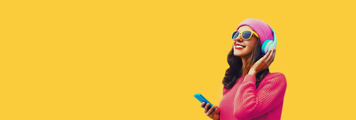 Portrait of happy smiling modern young woman in wireless headphones listening to music with smartphone wearing knitted sweater, pink hat on yellow background, blank copy space for advertising text