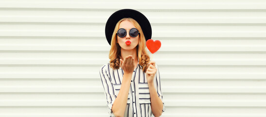 Portrait of beautiful young woman with red heart shaped lollipop blowing her lips with lipstick...