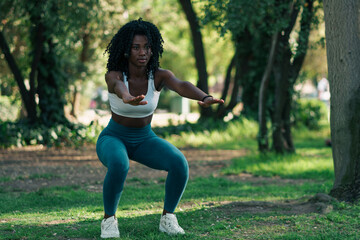 retrato de mujer negra con ropa deportiva, haciendo ejercicios de sentadilla, en un parque con luz...