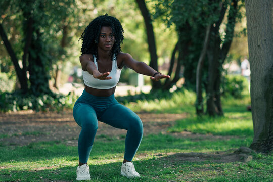 retrato de mujer negra con ropa deportiva, haciendo ejercicios de sentadilla, en un parque con luz natural. concentrada para hacer deporte