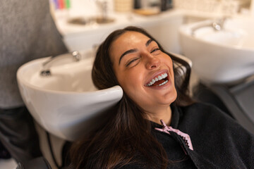 Delighted female client laughing and looking at camera while lying in sink during washing hair...