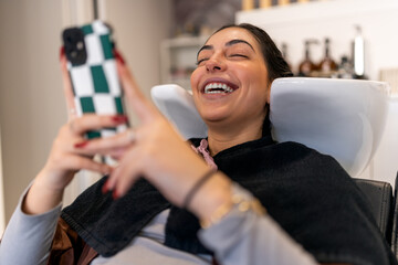 Positive young female smiling and browsing smartphone while lying in sink with closed eyes during beauty procedure in hairdressing salon
