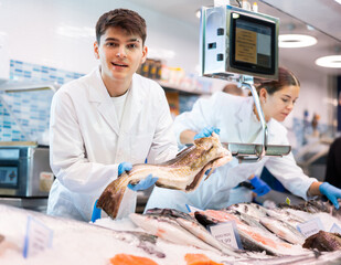 Positive young salesman demonstrating cod fish behind counter in fish store