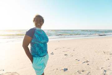 Fototapeta na wymiar Happy african american senior woman with short hair standing at sandy beach against sea and sky