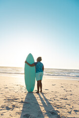 Fototapeta na wymiar Rear view of african american senior woman with surfboard looking at sea under blue sky at sunset