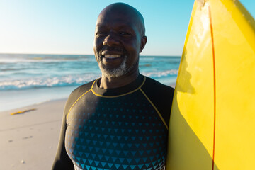 Smiling bald african american senior man with yellow surfboard standing against sea and clear sky