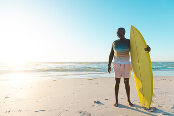 Fototapeta na wymiar African american senior man with yellow surfboard standing on sandy beach against sea and clear sky