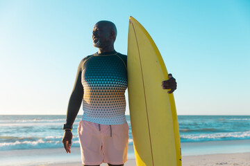 African american senior man with surfboard looking away while standing against sea and clear sky