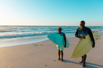 African american senior couple carrying surfboards while walking at sandy beach against sky