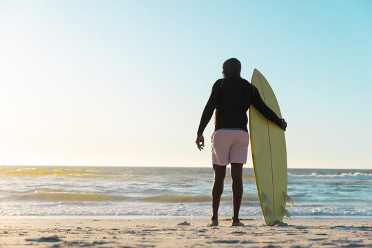 Rear View Of African American Senior Man With Surfboard Looking At Seascape And Clear Sky At Sunset