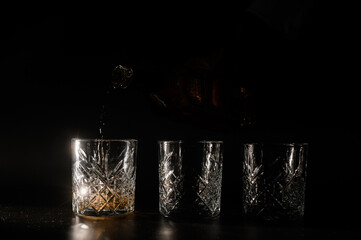 Front view of silhouette of hand with whiskey bottle pouring alcohol into set of crystal glasses, which placed on black background