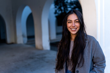 Positive young female in warm coat smiling and looking at camera while standing on pavement against arched passages of building in evening