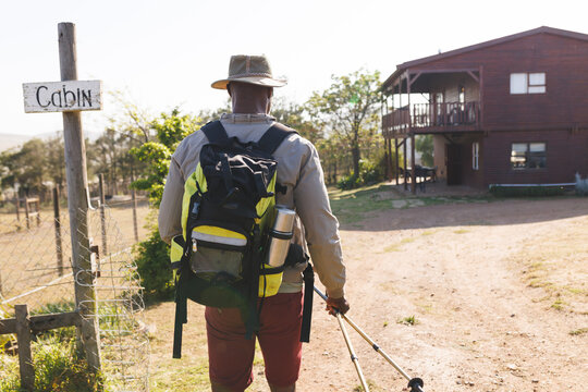 Rear view of senior african american man with hiking poles and backpack walking towards log cabin