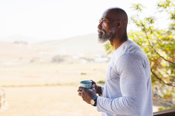 Bald african american senior man with mug contemplating and standing in balcony against clear sky