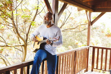 Bald african american senior man playing guitar and singing while sitting on railing in balcony