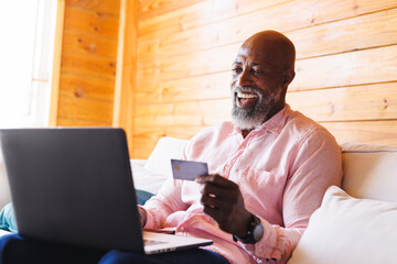 Bald african american senior man with credit card doing online shopping over laptop in log cabin