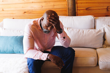 Stressed bald african american senior man with head in hand sitting on sofa in log cabin