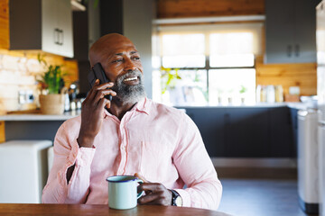 Smiling african american senior man holding coffee mug talking over smartphone in log cabin