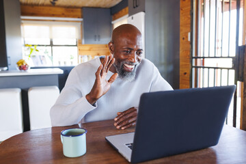 Happy african american senior man waving hand while talking over video call on laptop in log cabin