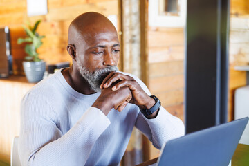 Serious bald african american senior man with hands clasped looking at laptop in log cabin