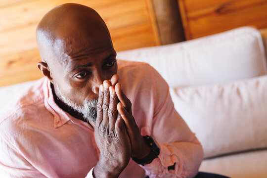 Close-up of worried bald african american senior man with hands clasped sitting on sofa in log cabin