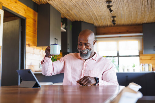 Bald african american senior man showing coffee mug over video call on digital tablet in log cabin