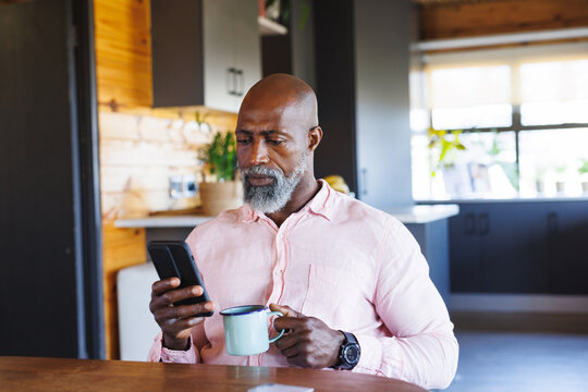 African american senior man with coffee mug and using smartphone while sitting at table in log cabin - Powered by Adobe