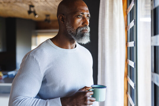 Thoughtful bald african american senior man with coffee mug looking through window at log cabin