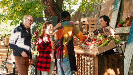 Diverse team of sellers helping customers choose natural products, selling homegrown eco fruits and vegetables. Local vendor showing bio fresh organic products at street greenmarket. Tripod shot.