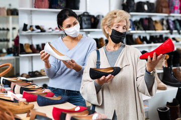 Fototapeta na wymiar Woman sales consultant in a protective mask, working in a shoe store during the pandemic, helps a mature european woman ..buyer choose ballet flats