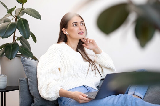 A young woman sits thoughtfully on a sofa with a laptop on her lap.
