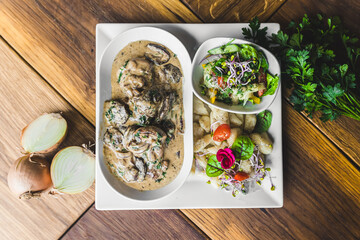 Top-down view of pork tenders in bolet mushroom sauce served on white plate with potato dumplings and fresh salad on rustic wooden table. Horizontal indoor shot. High quality photo