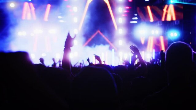 People clap their hands and dance at evening concert with light show.Fans raise their hands in bright colorful strobe lights on stage. Crowd is dancing, filming performance rock band, throwing party.
