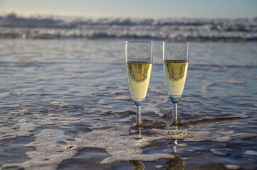 Glasses of cava or champagne sparkling wine on white sandy ocean beach with water waves on sunset in sunlights