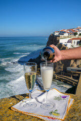 Pouring of champagne sparkling wine into glasses with view on white houses of picturesque village Azenhas do mar, Lisbon area, Portugal