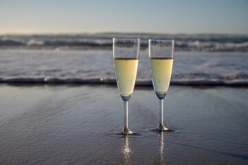 Glasses of cava or champagne sparkling wine on white sandy ocean beach with water waves on sunset in sunlights
