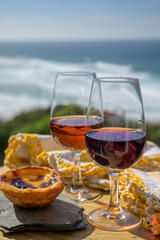 Portugal's food and drink, glass of porto wine, muscatel de setubal and sweet dessert Pastel de nata egg custard tart pastry served with view on blue Atlantic ocean in Lisbon area, Portugal