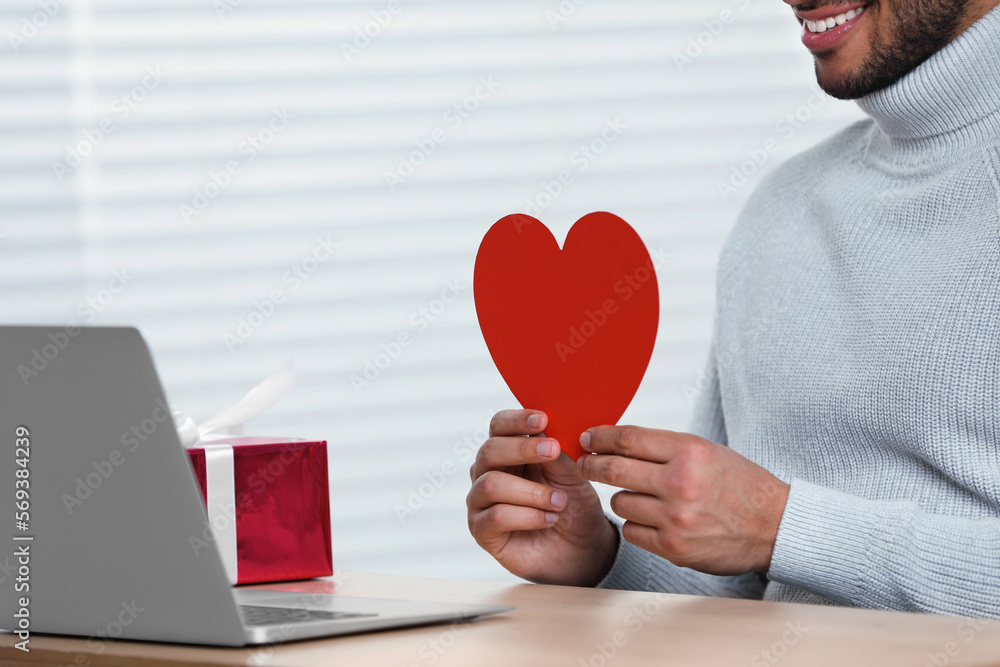 Poster Valentine's day celebration in long distance relationship. Man holding red paper heart while having video chat with his girlfriend via laptop, closeup