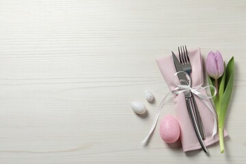 Cutlery set, painted eggs and beautiful flower on white wooden table, flat lay with space for text. Easter celebration