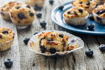 A blueberry muffin with the paper liner peeled back and several in behind.