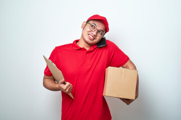 Smiling busy asian courier man answering phone call, holding mobile on shoulder, prepare package for delivery to client and making notes