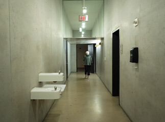 Eerie photo of a generic concrete bathroom hallway with drinking fountains and older woman walking out the door.