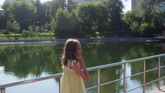 Happy smiling teen girl is walking near river in city calling on mobile phone. She is talking on smartphone on summer warm day walking alone down embankment. Child vacations, cellphone using concept.