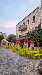 Greek Island Charm: Cozy Restaurant with colorful tables and chairs in front of Stone House, Colorful Architecture, and Lush Greenery	