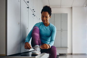Happy black athlete preparing for sports training and tying her shoelace in dressing room at gym.