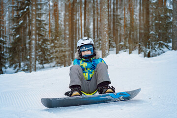 Fototapeta na wymiar little boy sitting on snow putting his feet in snowboard bindings adjusting straps.