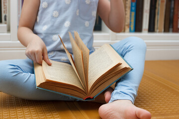 Little kid sitting in the front of bookshelf and holds few books in the hands. Concept of education