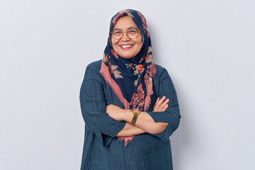 Elderly smiling Asian Muslim woman 50s wearing hijab in glasses crossed arms and looking confident...