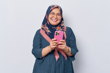 Elderly smiling Asian Muslim woman 50s wearing hijab in glasses holding smartphone and smiling at camera isolated on white background. People islam religious concept